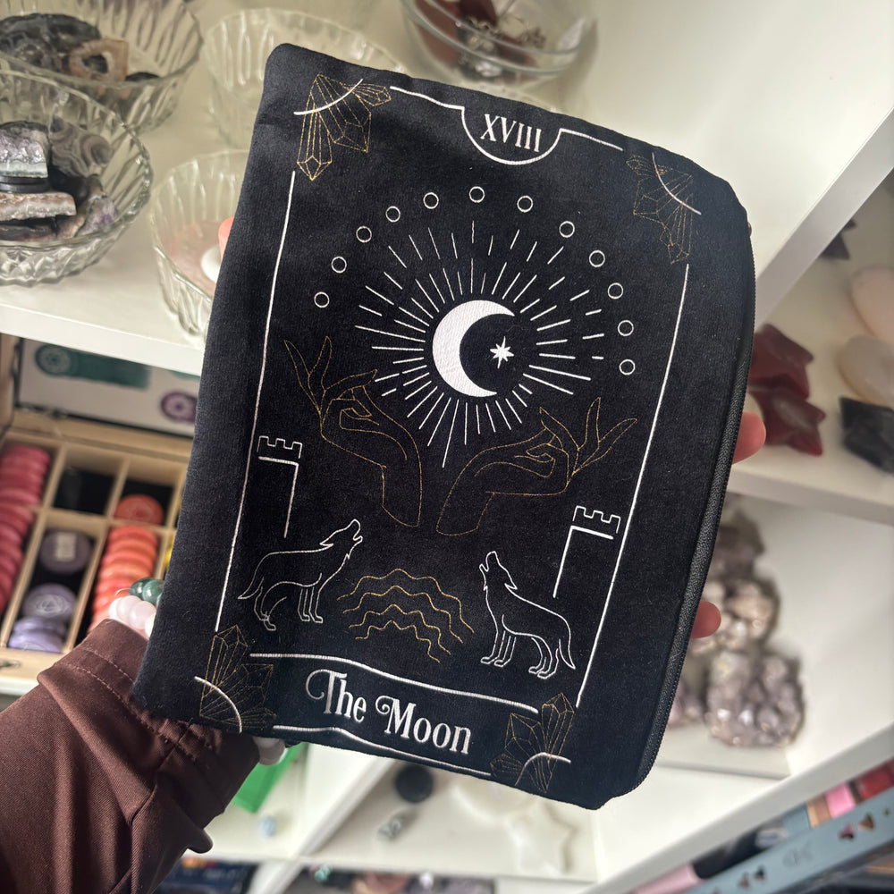 A hand holding a black velvet oracle card deck holder bag in front of a white shelf filled with crystals