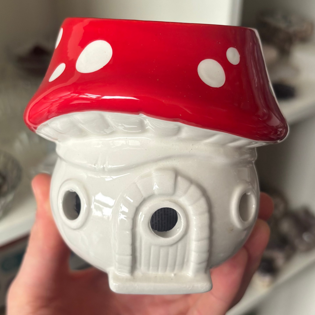 A close-up view of a whimsical mushroom-shaped ceramic oil burner held upright in a hand. Designed as a fairytale cottage, the smooth white ceramic base features small circular window cut-outs beneath a glossy, bright red toadstool cap detailed with bold white spots.