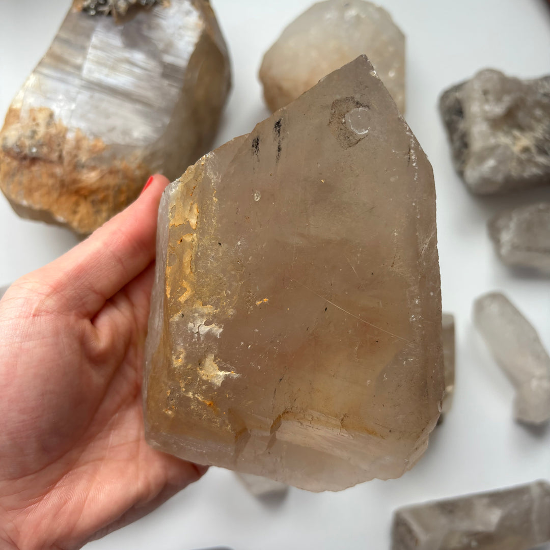 A hand displaying the wide base and textured, chunky contours of a geological raw quartz crystal piece, illustrating the impressive size of this crystalline powerhouse used to naturally cleanse and recharge an entire collection.