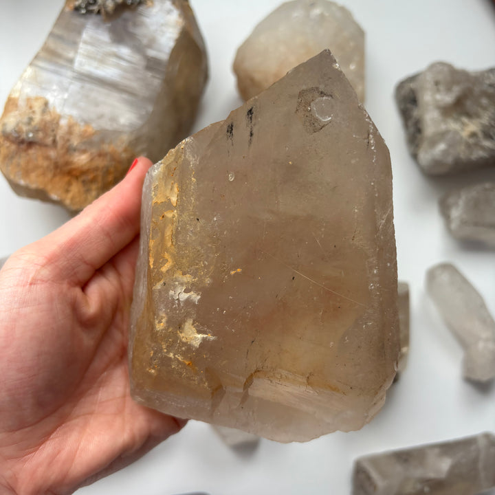 A hand displaying the wide base and textured, chunky contours of a geological raw quartz crystal piece, illustrating the impressive size of this crystalline powerhouse used to naturally cleanse and recharge an entire collection.