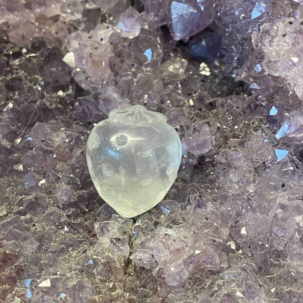 A wide shot looking down at the carved quartz strawberry crystal sitting gracefully amidst raw purple gemstone points. This layout emphasizes the small, two-and-a-half-centimetre scale, offering a sweet visual anchor for a desktop display tray.
