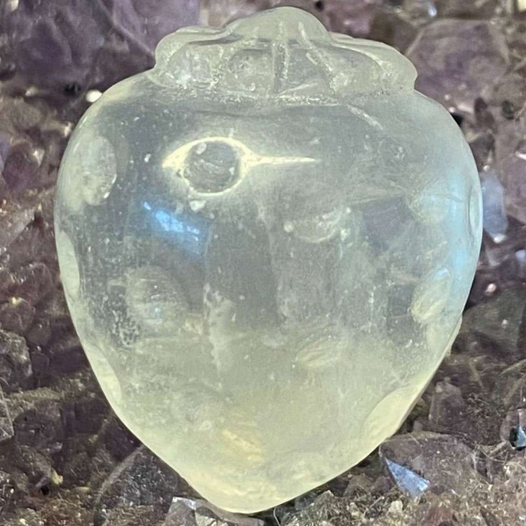 A close-up view of the small, polished Clear Quartz Strawberry Carving. The translucent fruit figurine showcases glass-like textures and tiny, frosty internal birthmarks, resting on a sparkling bed of purple amethyst druzy points.