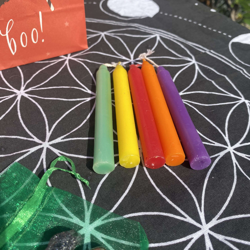 A close-up of five slim, vibrant ritual chime candles next to a green organza pouch on a geometric cloth.