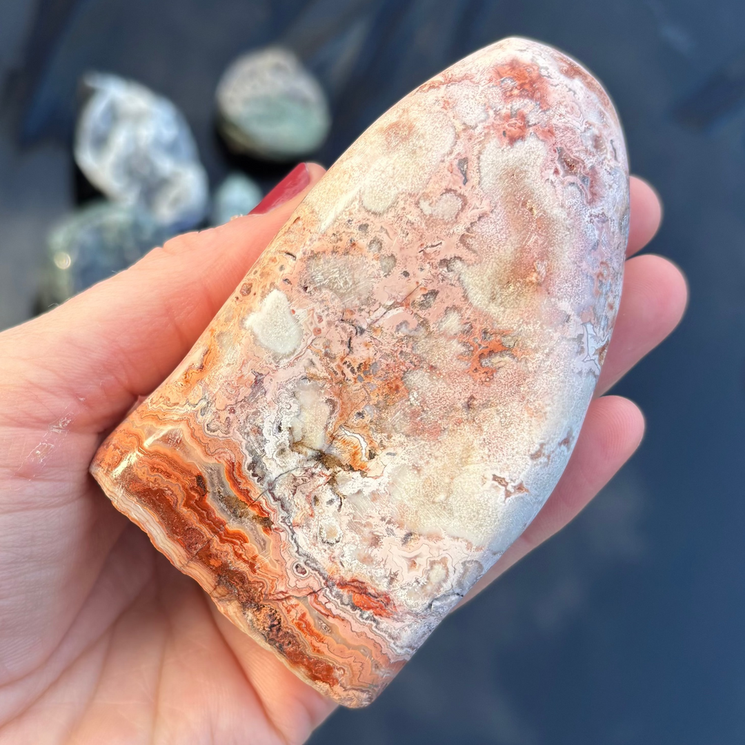 A polished Cotton Candy Agate freeform crystal cradled in an open palm, showcasing its smooth glossy face woven with swirling pastel pink, cream, and apricot ribbons outdoors.
