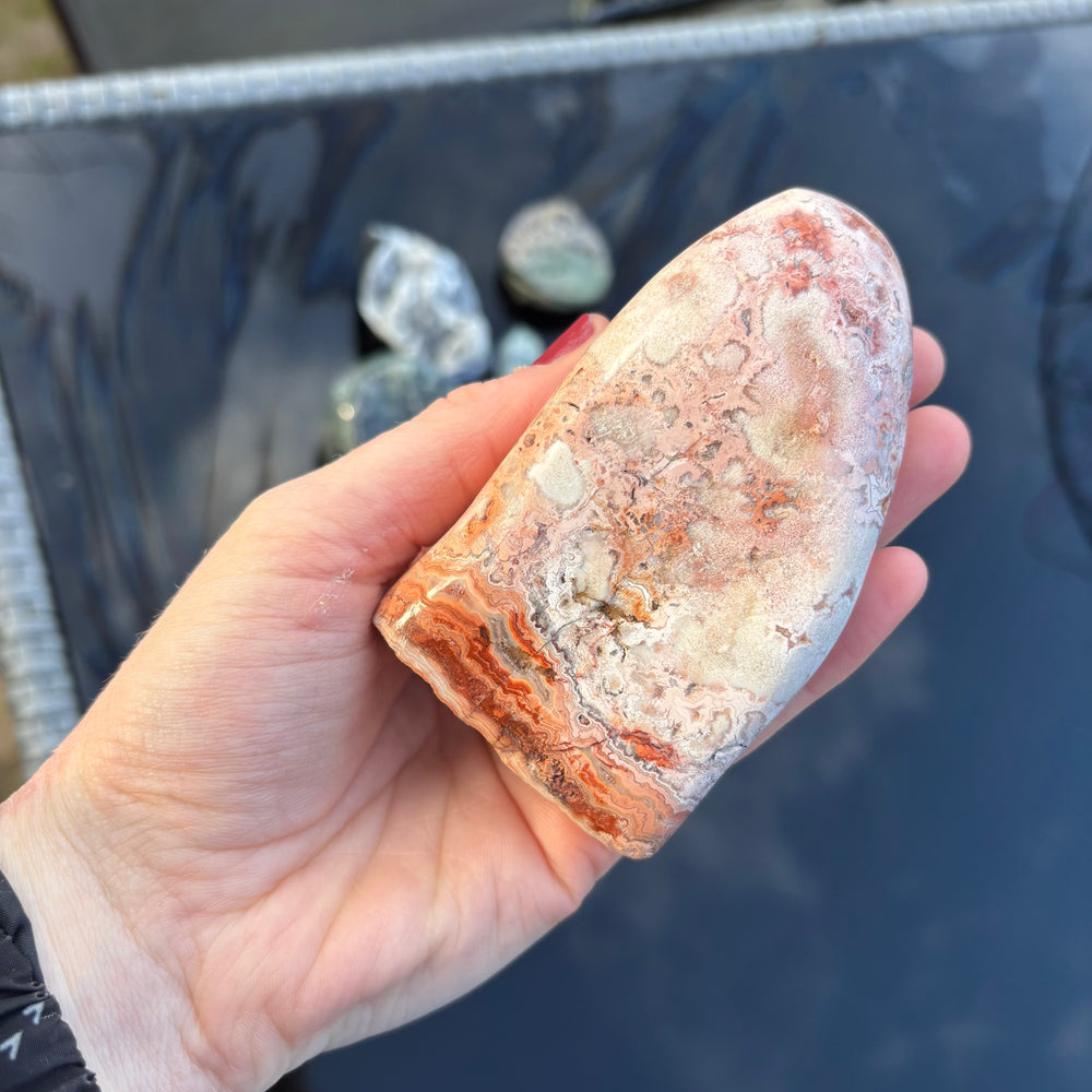 A hand holding the sculpted Cotton Candy Agate freeform stone upright from a side profile angle, highlighting its flat standing base and thick geological layers under bright daylight.