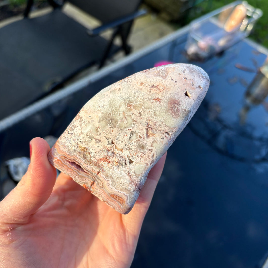 A hand presenting the thick underside and lower crystalline profile of the Cotton Candy Agate freeform outdoors, highlighting its sturdy polished depth and natural cream mineral textures.