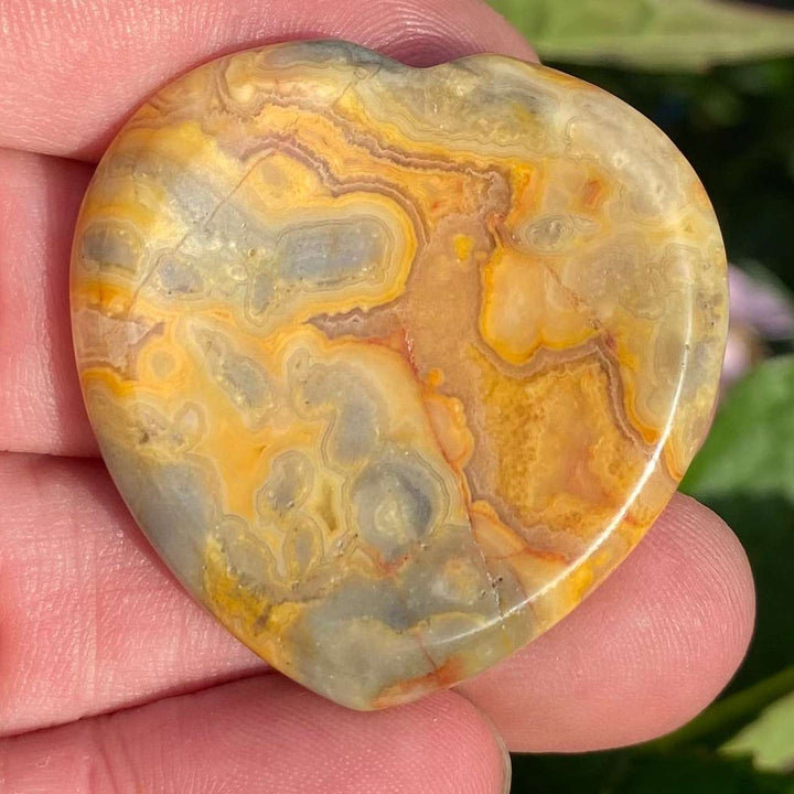 A close-up view of a polished Crazy Lace Agate Heart Worry Stone held flat in an open hand. The smooth crystal surface showcases marbled yellow, orange, gold, and grey patterns, serving as a tactile pocket companion to invite optimism, grounding energy, and solar plexus chakra alignment.