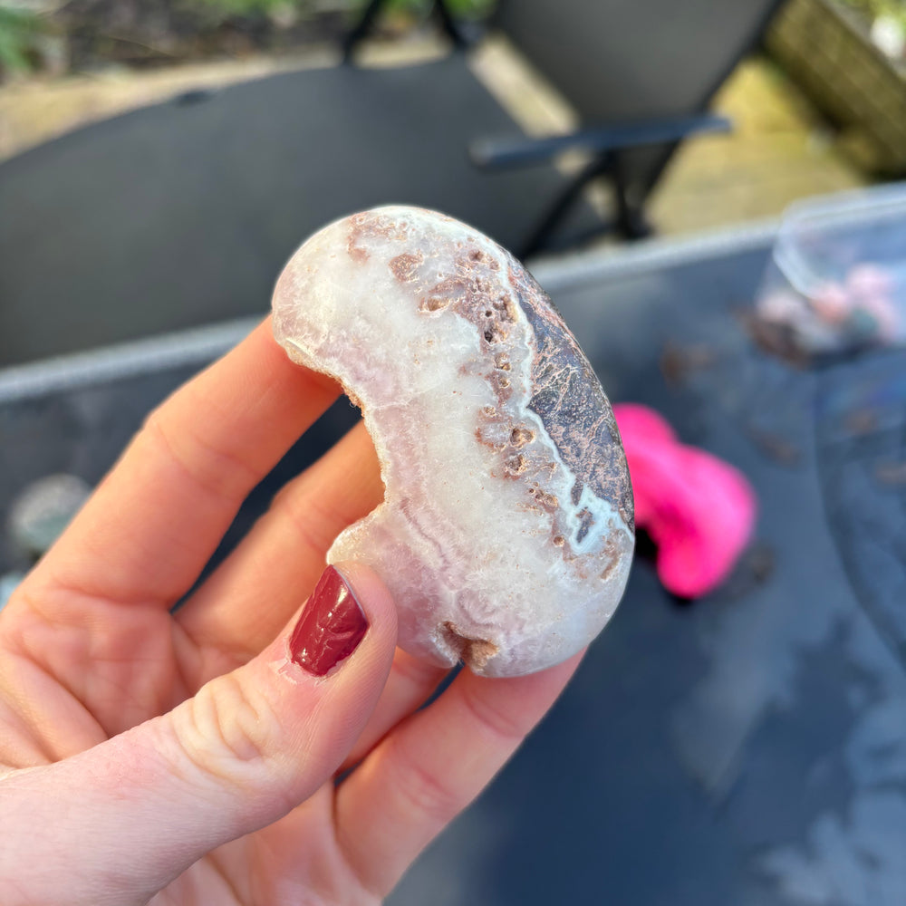 An angled overhead view looking down at the distinctive Pink Aragonite stone held carefully against a outdoor table backdrop. The natural morning light illuminates the smooth, velvety textures and organic brown-and-white banding, offering a comforting presence to steady scattered thoughts.