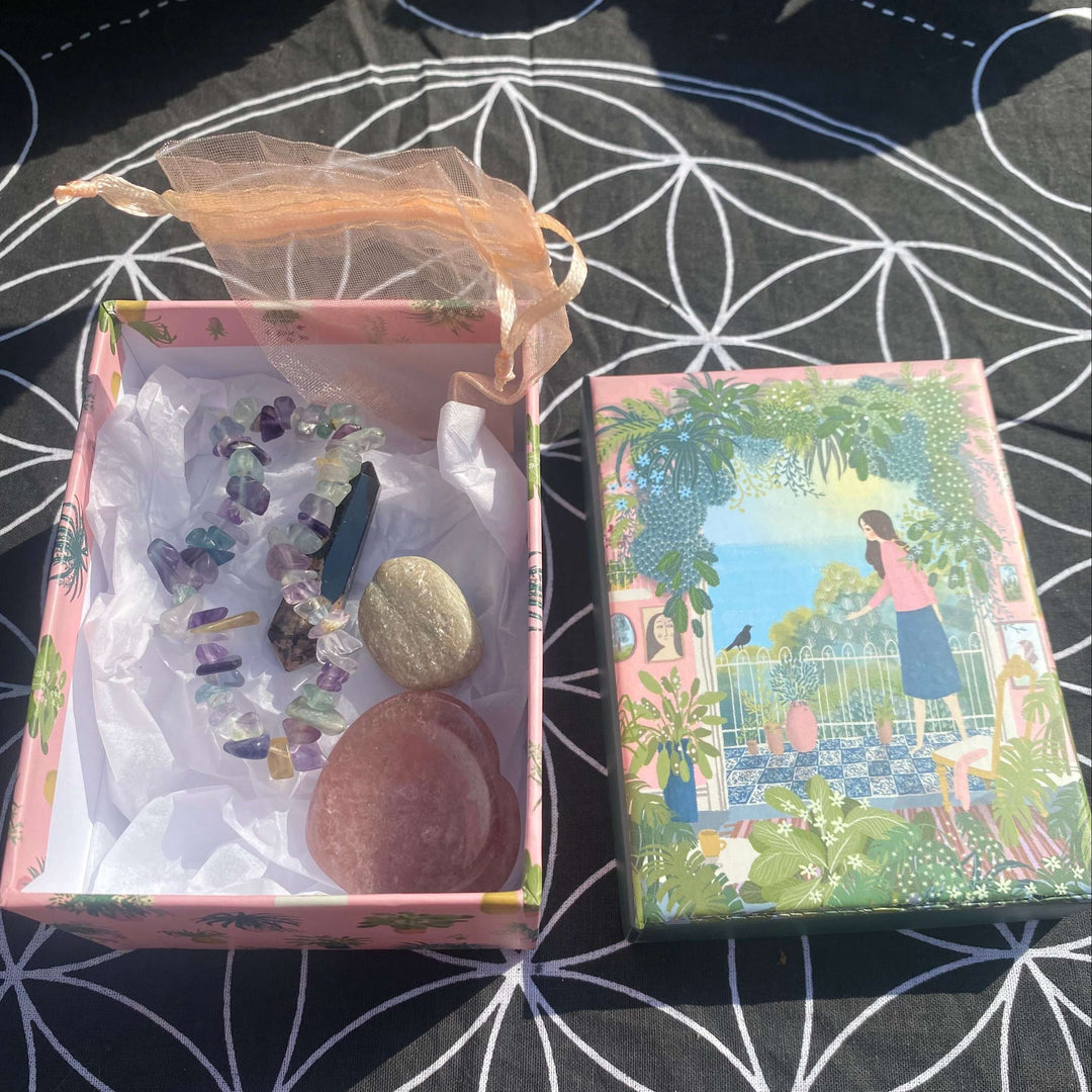 A complete crystal gift set displayed unboxed outdoors on a black sacred geometry mat. The patterned pink gift box is open, revealing a fluorite chip bracelet, a polished rhodonite generator point, a smooth aventurine heart, and an organza bag against a beautiful illustrated green lid.