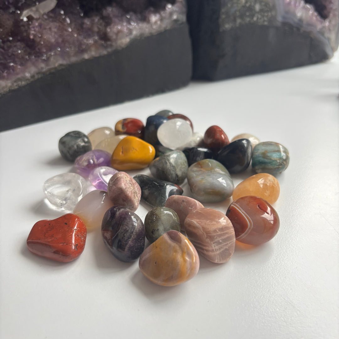 A clustered pile of smooth mystery tumble stones arranged on a white table layout, detailing the unique patterns, earthy shading variations, and minor chips of these discounted items styled in front of large amethyst crystal clusters.