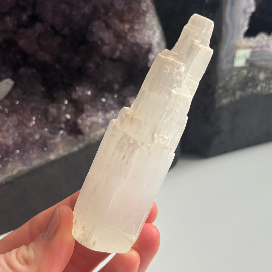 A hand holding the discounted satin spar selenite tower at an angle indoors, detailing the light-reflective mineral ridges, jagged skyscraper stepped tiers, and minor surface scratches that bring a soft frequency of calm serenity.