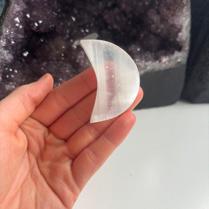 An open palm presenting the discounted selenite half moon pocket stone flat indoors, highlighting the vertical satin grains and unique natural surface scratches of this budget-friendly clearance crystal.