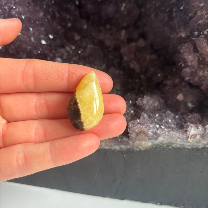 A hand presenting the reverse side pattern of the discounted septarian jasper tear drop carving indoors, detailing the prominent golden coloration and a small physical chip right on its pointed tip to show its unique character.