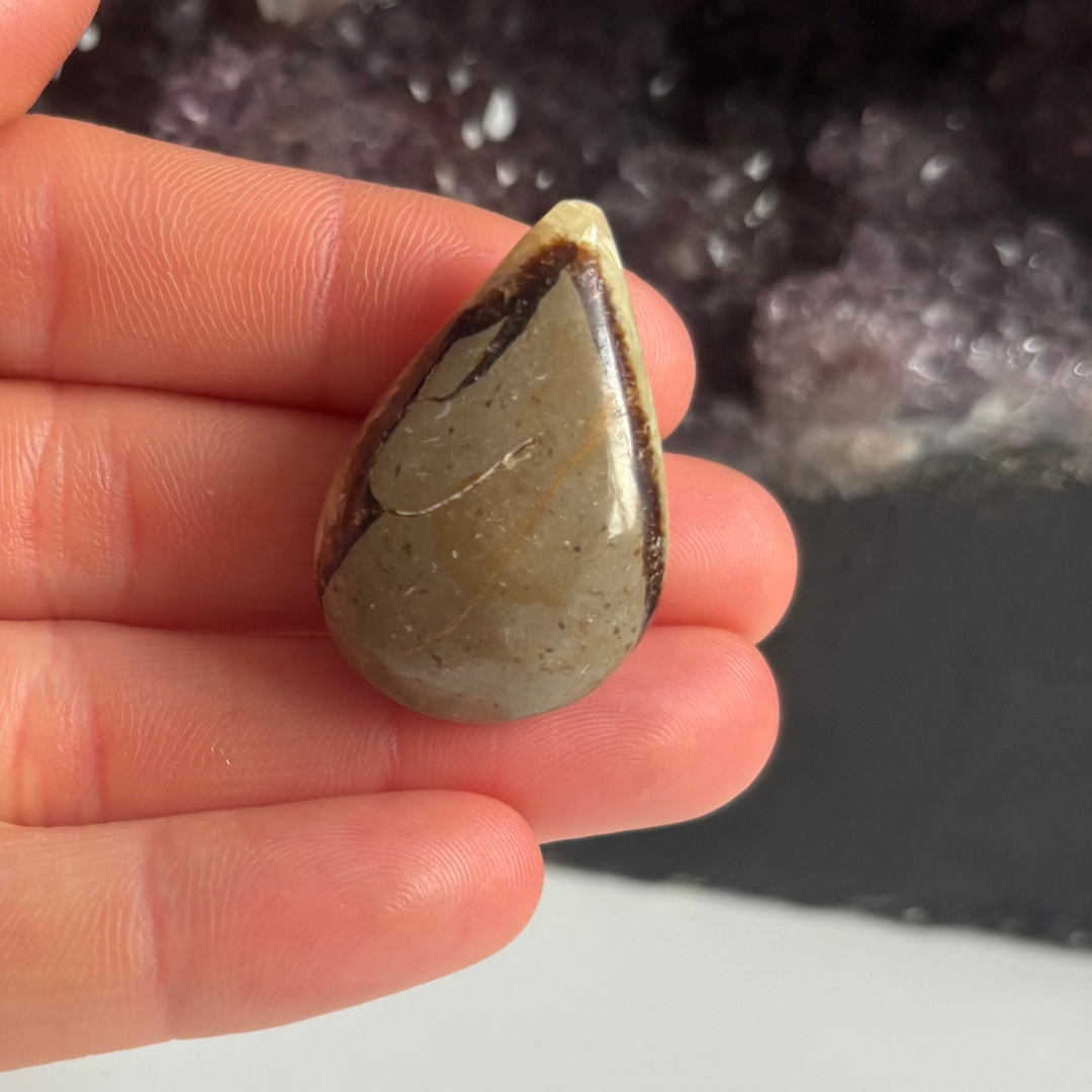 A hand holding the reverse side of the discounted septarian jasper tear drop carving, revealing a predominantly solid brownish-grey mineral surface and a small physical chip right on its pointed top area to show its clearance character.
