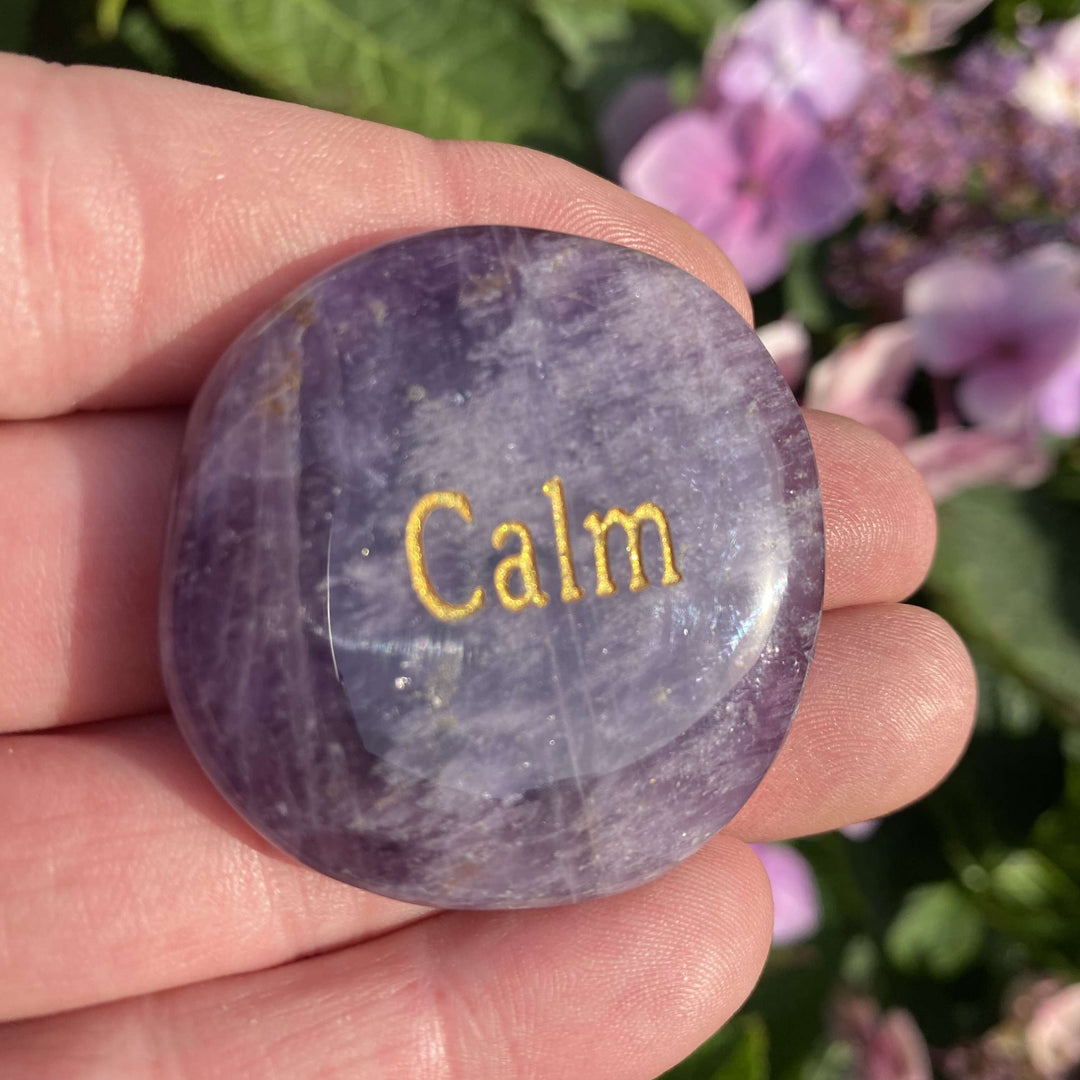 A close-up view of an oval dream amethyst palm stone held in an open hand, featuring the word Calm engraved in gold letters across a polished purple and white marbled surface with a background of pink garden flowers.
