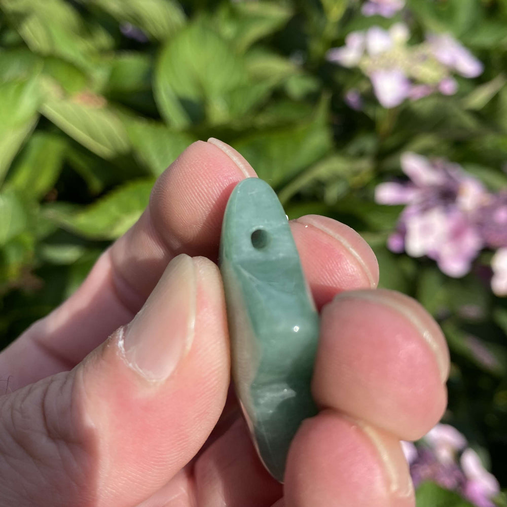A side profile view of the Green Aventurine star carving held between fingers outdoors, showcasing the small pre-drilled threading hole. The daylight highlights its compact thickness and smooth polished edge, ready to be threaded onto a necklace cord or ritual ribbon.