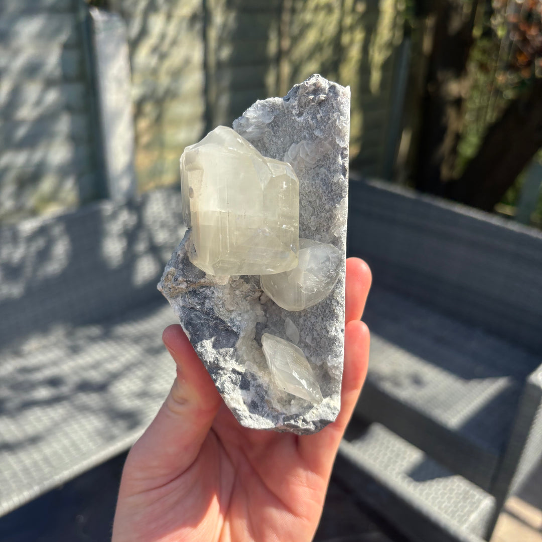 A hand holding the raw grey chalcedony and calcite crystal cluster vertically, demonstrating its earthy, unpolished side profile and substantial geological layers that bring emotional stability and reassurance to a personal collection.