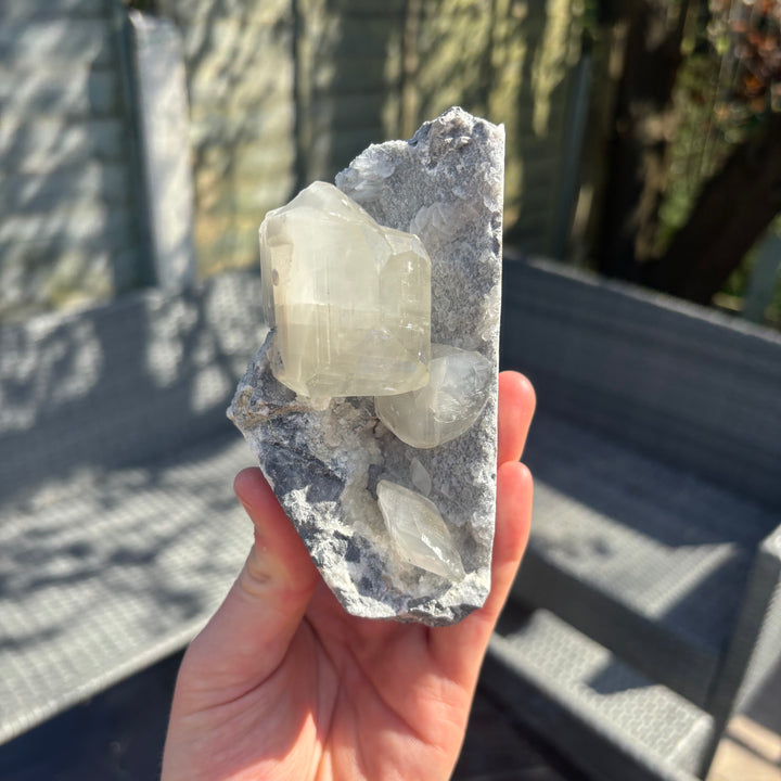 A hand holding the raw grey chalcedony and calcite crystal cluster vertically, demonstrating its earthy, unpolished side profile and substantial geological layers that bring emotional stability and reassurance to a personal collection.