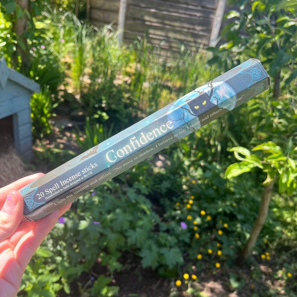 A hand holding the thin hexagonal pack of black confidence spell incense sticks at a distance outdoors, highlighting the sweet and floral musk fragrance companion styled against green garden leaves to bring comforting warmth.