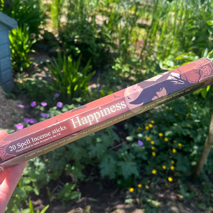 A hand holding a brown and golden-yellow hexagonal pack of Elements Happiness Spell incense sticks close up, featuring mystical artwork by Lisa Parker against a lush, sunlit garden backdrop filled with green foliage and flowers.
