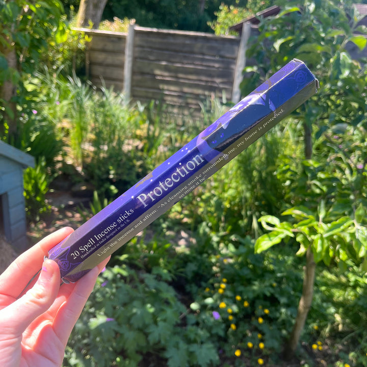 A hand holding the thin hexagonal pack of blue protection spell incense sticks at a distance, showcasing the woody sandalwood fragrance companion styled against sunlit garden greenery to welcome a comforting, grounding energy and deep security into the home.