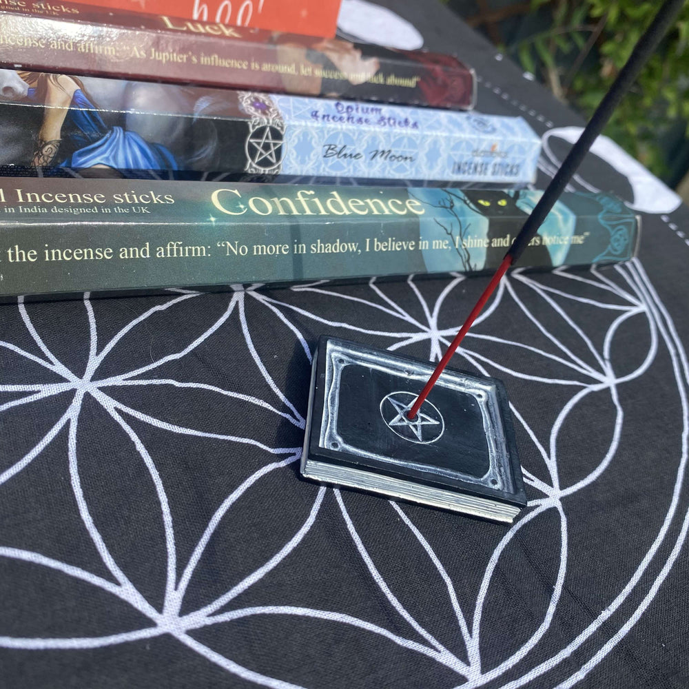 A detailed view looking down at a burning incense stick resting in a small square pentacle holder. In the softly blurred background, the colourful ritual packaging for the Confidence and Luck spell incense boxes sits on a patterned altar cloth under bright daylight.