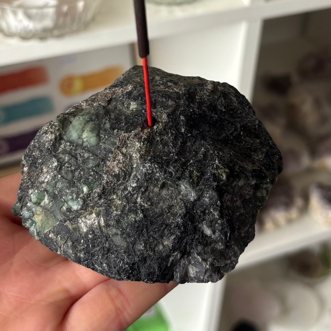 A close-up view of an open hand supporting a heavy, raw emerald crystal incense stick holder with an incense stick inserted, showcasing dark green colour zones and natural texturised fractures indoors against a blurred white shelving unit background for Heart Chakra alignment.