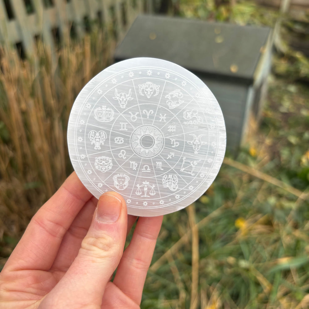A hand presenting the engraved circular selenite charging disk at a distance outdoors over a garden setting, demonstrating the approximate 8cm sizing scale and translucent pearlescent sheen of this cosmic cleansing tool.