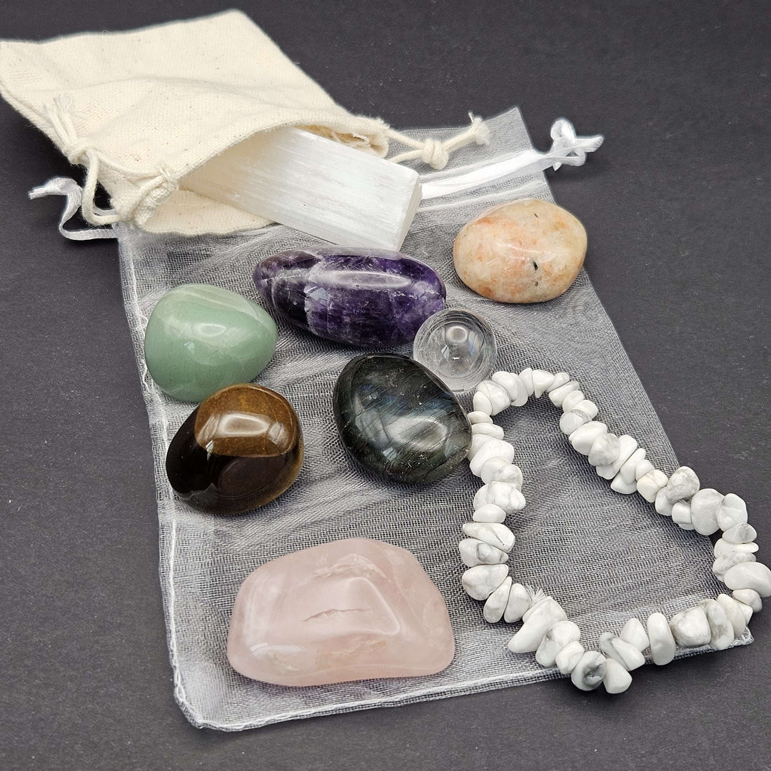 A close-up lifestyle view of the Everyday Bracelet and Crystal Set displaying a white Howlite chip stretch bracelet. The beaded jewellery rests over a grey organza storage bag next to polished amethyst, rose quartz, and tiger eye tumbled stones. 