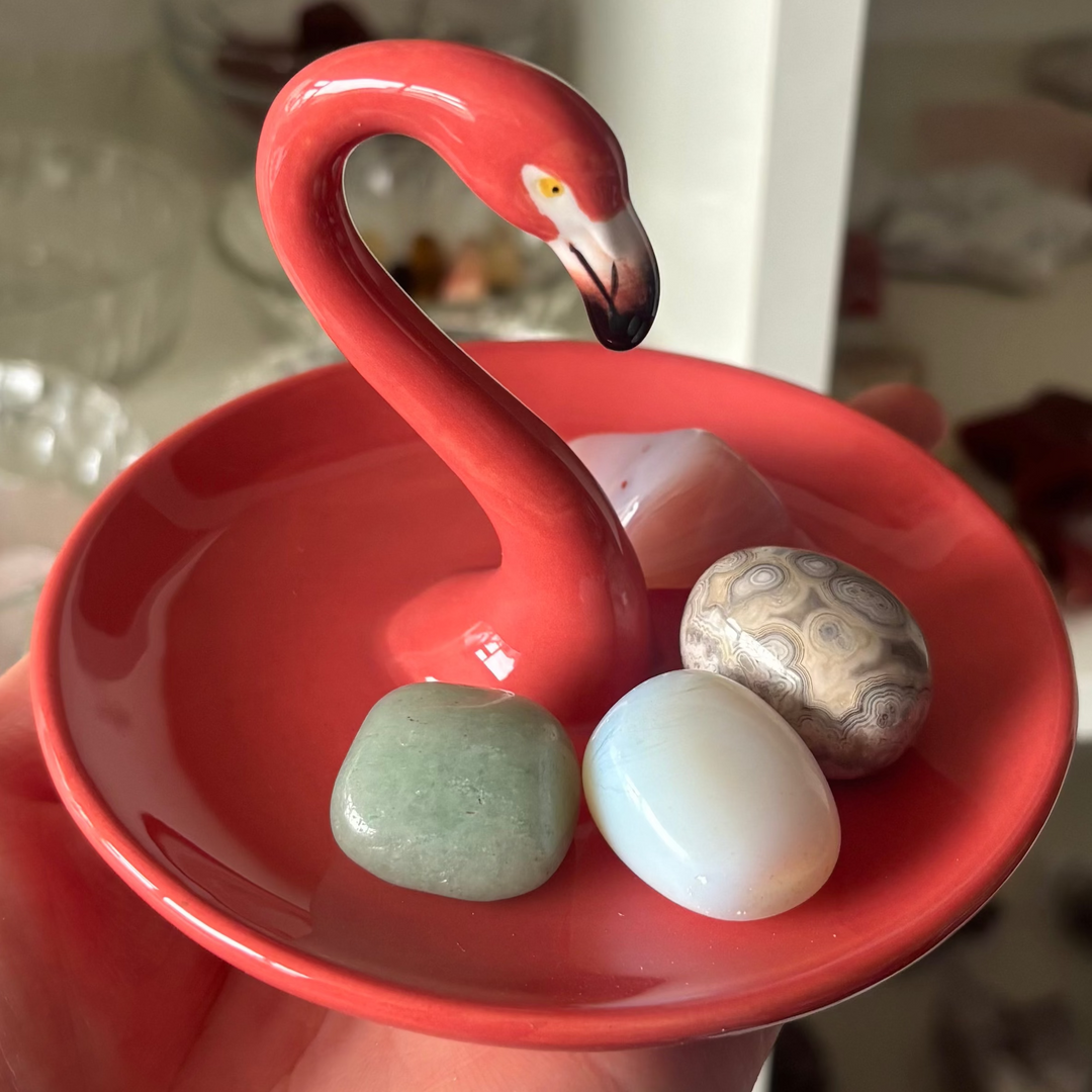 A close-up view of the ceramic Flamingo Ring Holder held in a hand, showcasing its smooth glossy finish and vibrant coral pink glaze. The round base dish securely cradles three polished crystal tumble stones, highlighting a playful magic and heart chakra sanctuary vibe.