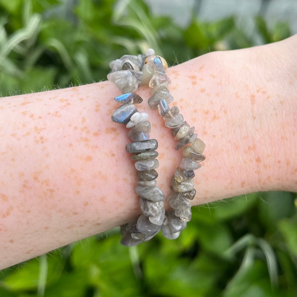 A mid-range outdoor view of a wrist showcasing two stacked labradorite gemstone chip bracelets against a fresh background of green garden foliage, displaying the organic, bumpy stone textures and flexible 18cm stretchy styling scale.