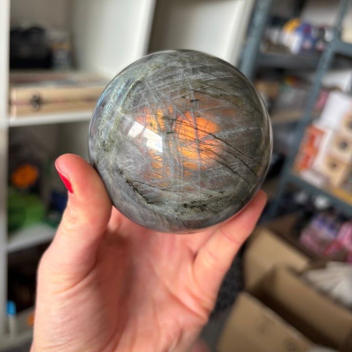 A hand tilting the flashy labradorite orb to reveal a brilliant fiery orange and bronze streak radiating from its centre, demonstrating the premium iridescence of this 696-gram mystical statement piece.