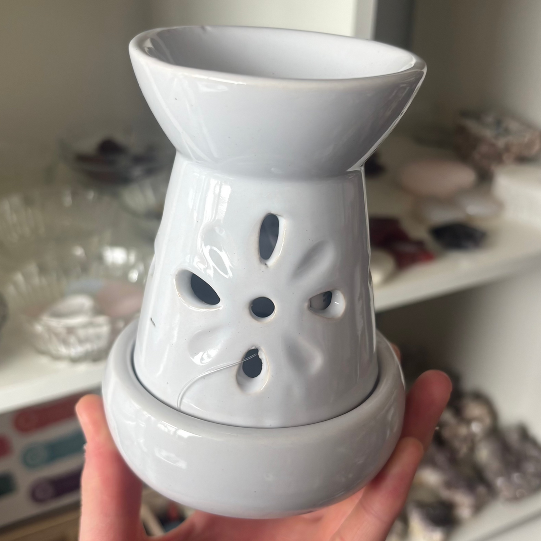 A close-up view of the smooth white ceramic Floral Cut Out Oil and Wax Burner held upright in an open hand. The botanical design features delicate cut-out petal patterns, casting a pretty candlelight glow to enhance a cosy room atmosphere and align your heart chakra.
