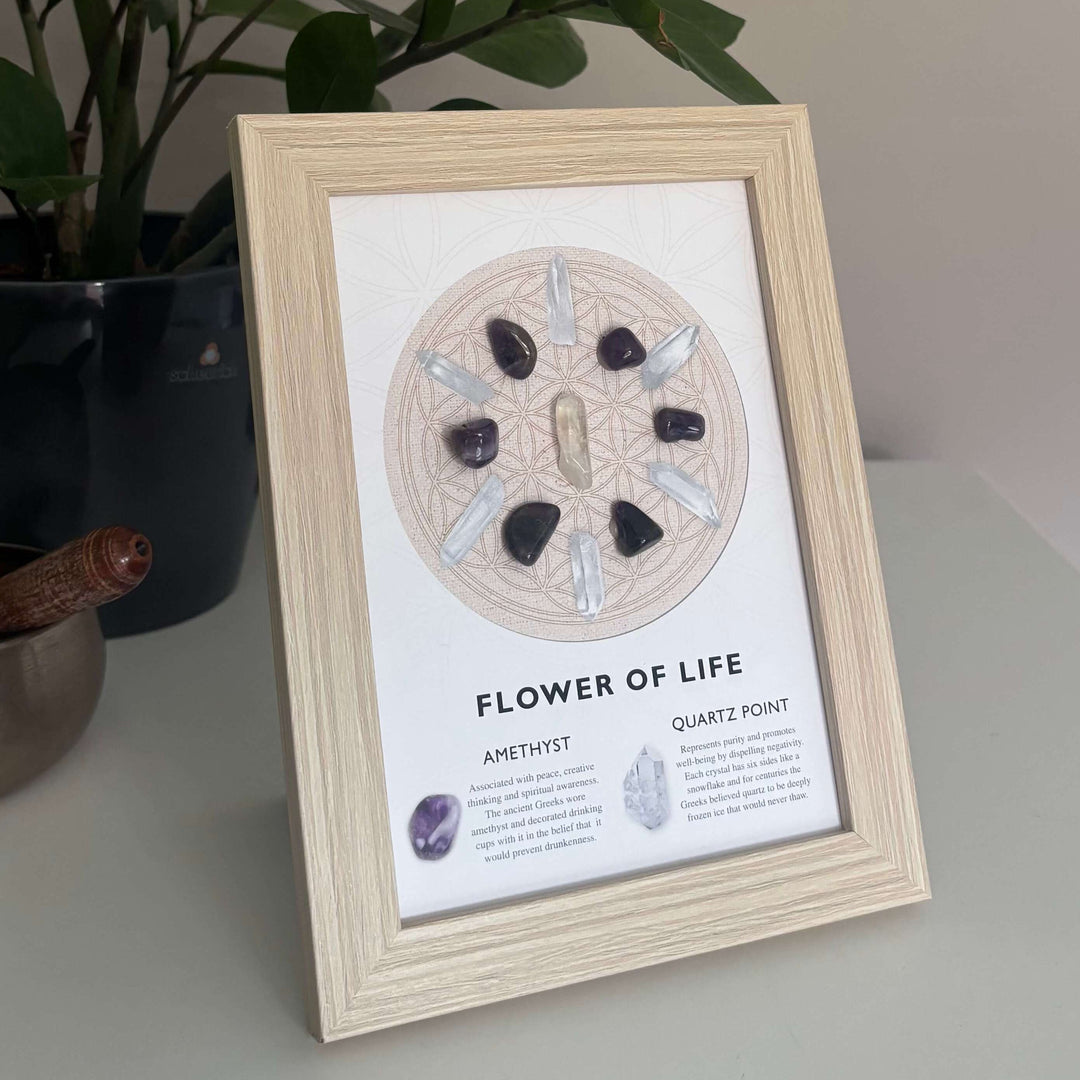 A close-up view of a light-coloured wooden frame displaying the Flower of Life crystal artwork upright on a table. The sacred geometry design features real amethyst and clear quartz crystal pieces arranged symmetrically, styled beautifully next to a dark potted houseplant to highlight its Crown Chakra alignment.
