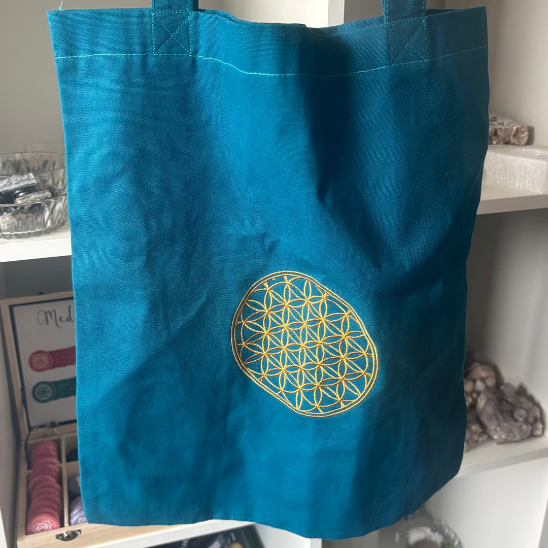 A front-on close-up shot of a teal blue canvas tote bag hanging inside a boutique crystal shop. The lower right portion of the textured cotton shopper features an intricate, circular geometric Flower of Life pattern stitched in vibrant yellow embroidery thread, creating a mindful aesthetic against blurred white store shelves.