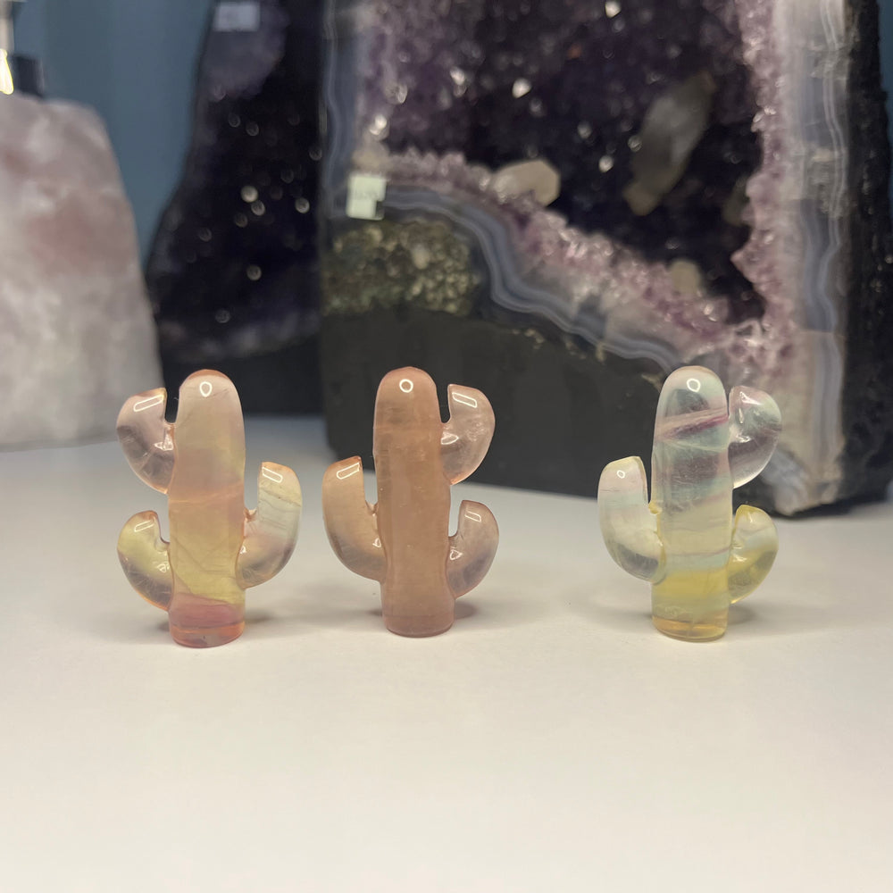 A wider shot of three miniature fluorite cactus crystal carvings standing in a row on a white table layout, demonstrating their whimsical botanical shapes that help sweep away mental clutter and bring a delightful vibe to a workspace desk.
