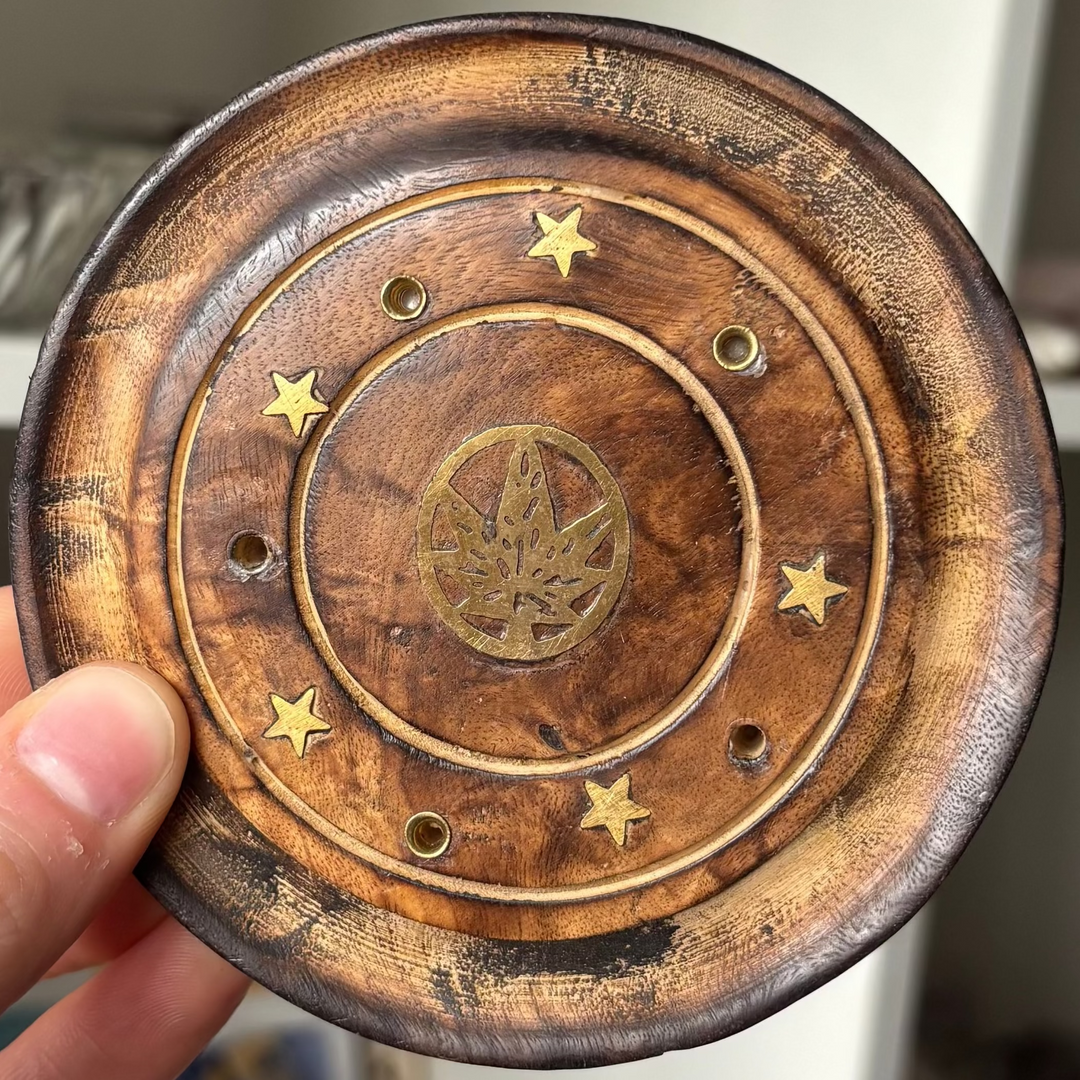 A close-up view of the round gold leaf incense stick holder held gently in a hand. The design showcases the natural mango wood with concentric circular grooves, small star-shaped brass inlays, and a detailed central leaf emblem to channel abundance and clarity.