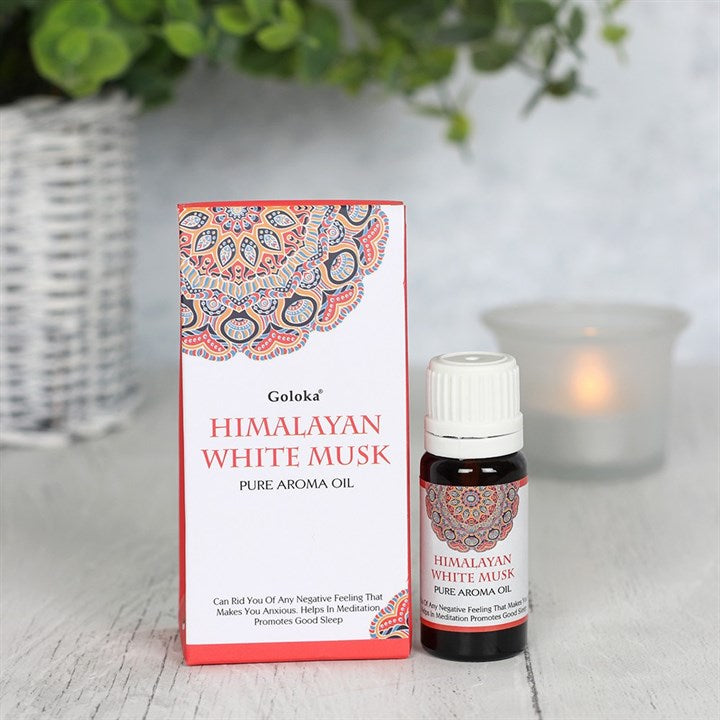 A 10ml bottle of Goloka Himalayan White Musk pure aroma oil standing next to its red and white decorative mandala card box packaging, styled indoors on a light tabletop in front of a glowing frosted glass candle burner dish.