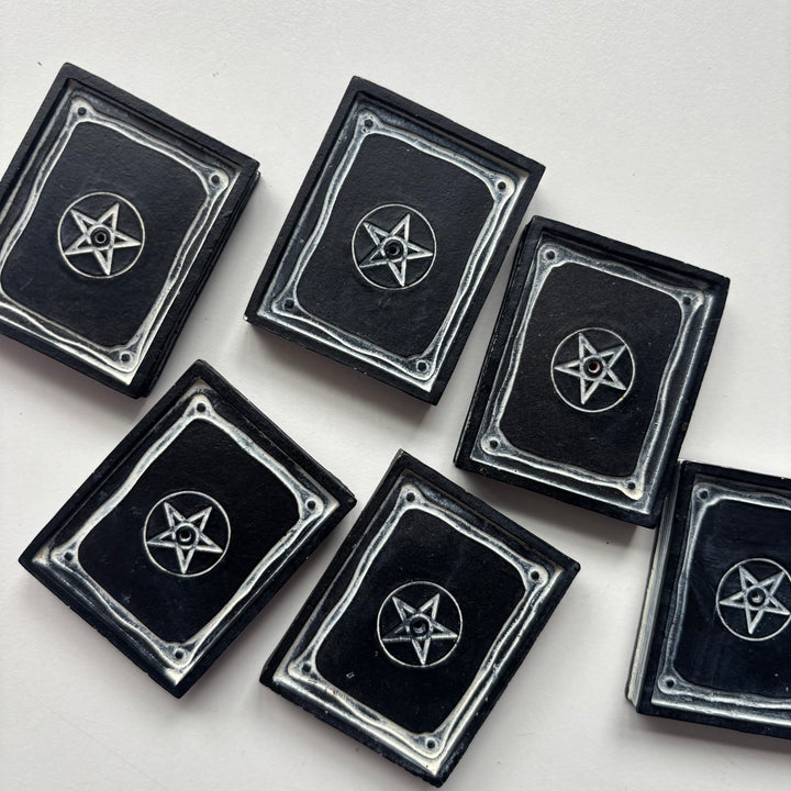 An overhead shot looking down at a collection of small black spell book incense plates scattered across a smooth white surface. The clean indoor lighting highlights the crisp silver pentagram designs and raised book borders, demonstrating their compact real-world scale and whimsical styling potential.