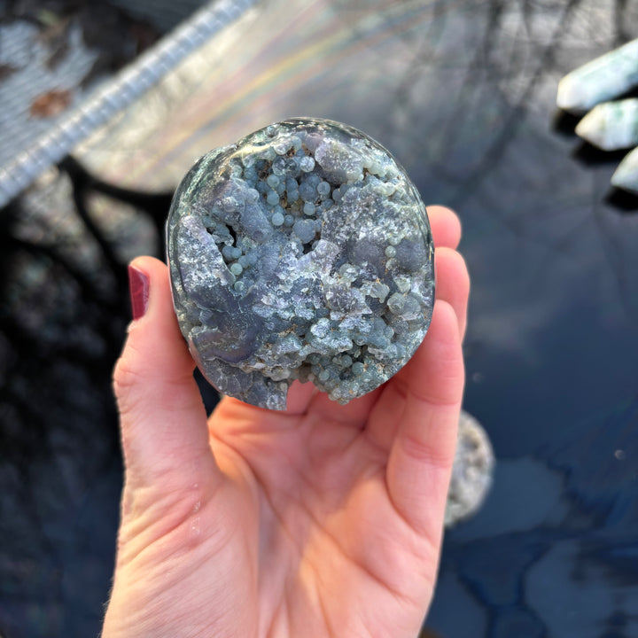 A hand presenting a round, raw Grape Agate crystal freeform to illustrate its comfortable, pocket-sized weight for quiet ritual moments.