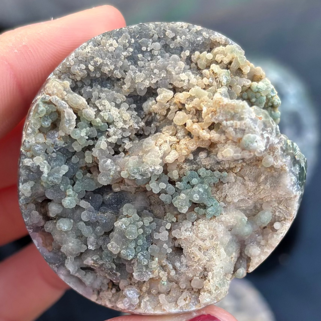 A close-up shot of a round Grape Agate crystal freeform held in a hand, showing its unique bubbly botryoidal cluster texture in shades of pale purple, grey, and green.