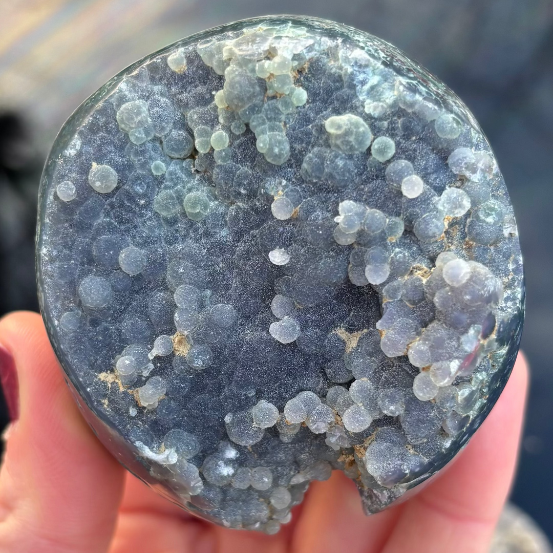 A close-up shot of a round Grape Agate crystal freeform held in a hand, showing its unique botryoidal texture with pale purple and green chalcedony spheres.