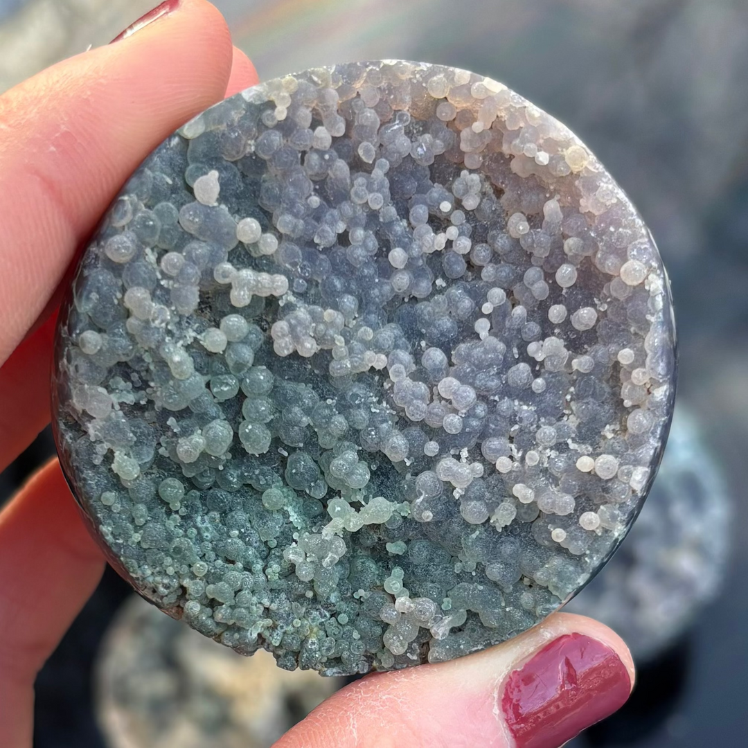 A round, polished Grape Agate crystal freeform held upright in fingertips, showcasing a textured face split between earthy matte green and a dense patch of tiny lavender and deep purple botryoidal spheres.