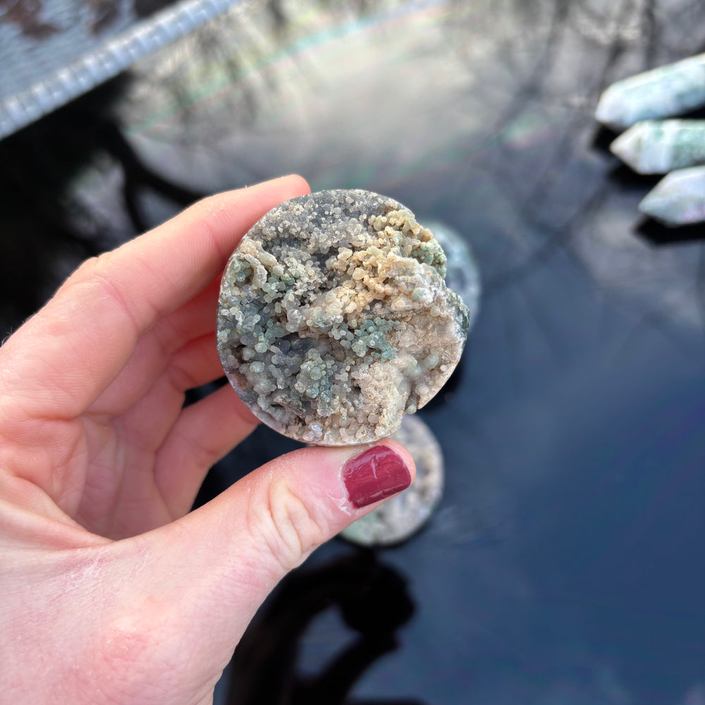 A hand holding a small, spherical polished Grape Agate stone over a dark reflective surface, showcasing its round shape and organic patterns.