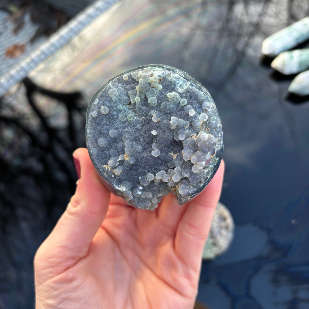 A hand holding a chunky, round Grape Agate freeform stone over a dark reflective surface, showcasing its organic shape and natural mineral layers.