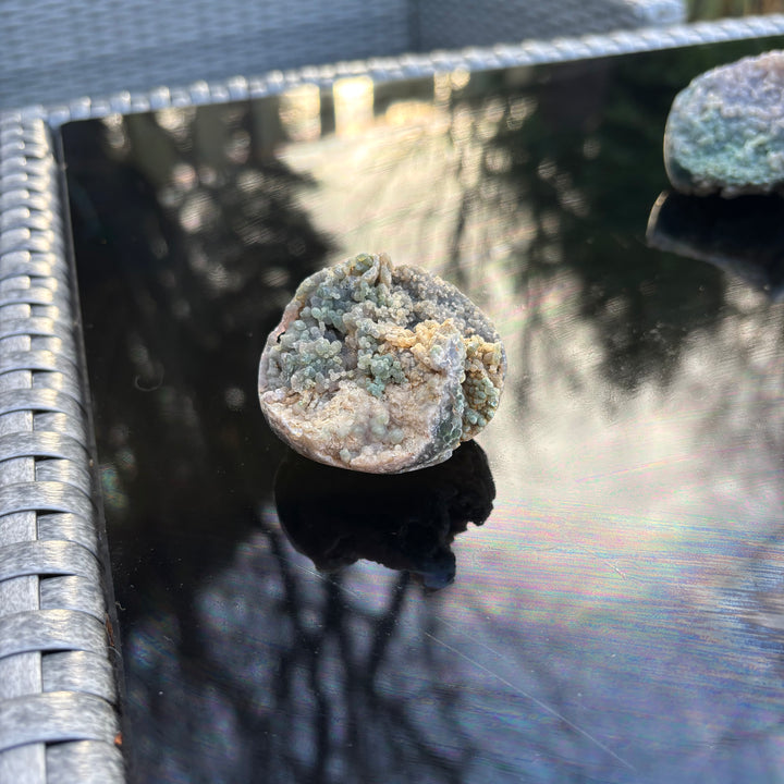 A round Grape Agate crystal specimen resting solo on a black glossy table, catching natural outdoor light and creating a soft reflection.
