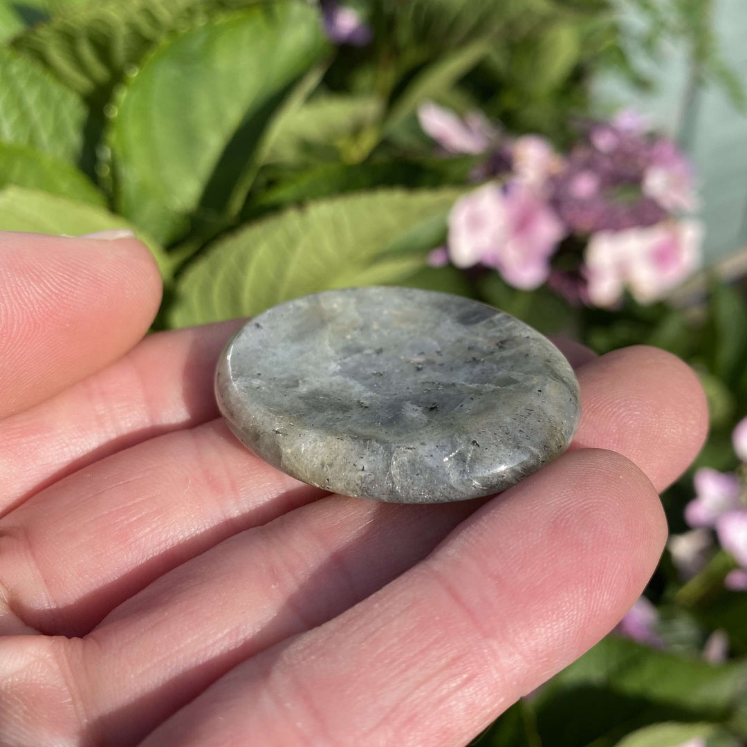 A close-up view of a dark gray Labradorite worry stone angled gently on an open hand in bright sunlight. The celestial sheen and subtle blue flashes showcase its untamed cosmic energy, making it an enchanting, thoughtful gift to support natural intuition and the Crown Chakra.
