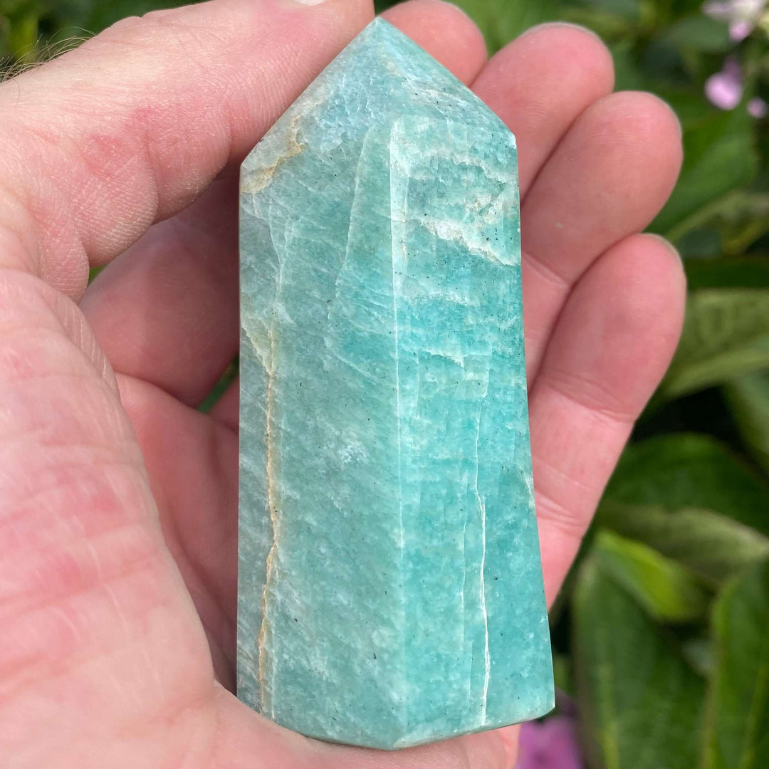A macro close-up view of a Green Amazonite crystal generator tower point held vertically in an open palm. The six-sided gemstone pillar displays a rich minty-blue to light sea-green hue with distinct, organic white streak inclusions across its smooth polished facets.