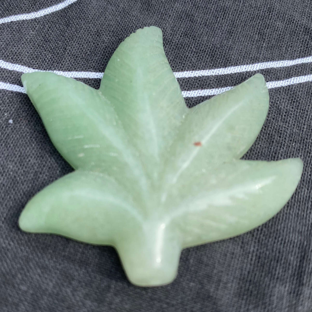 A close-up view of a pale green aventurine carved leaf crystal displaying a slight natural surface mark on a dark gray fabric backdrop with white woven stripes.
