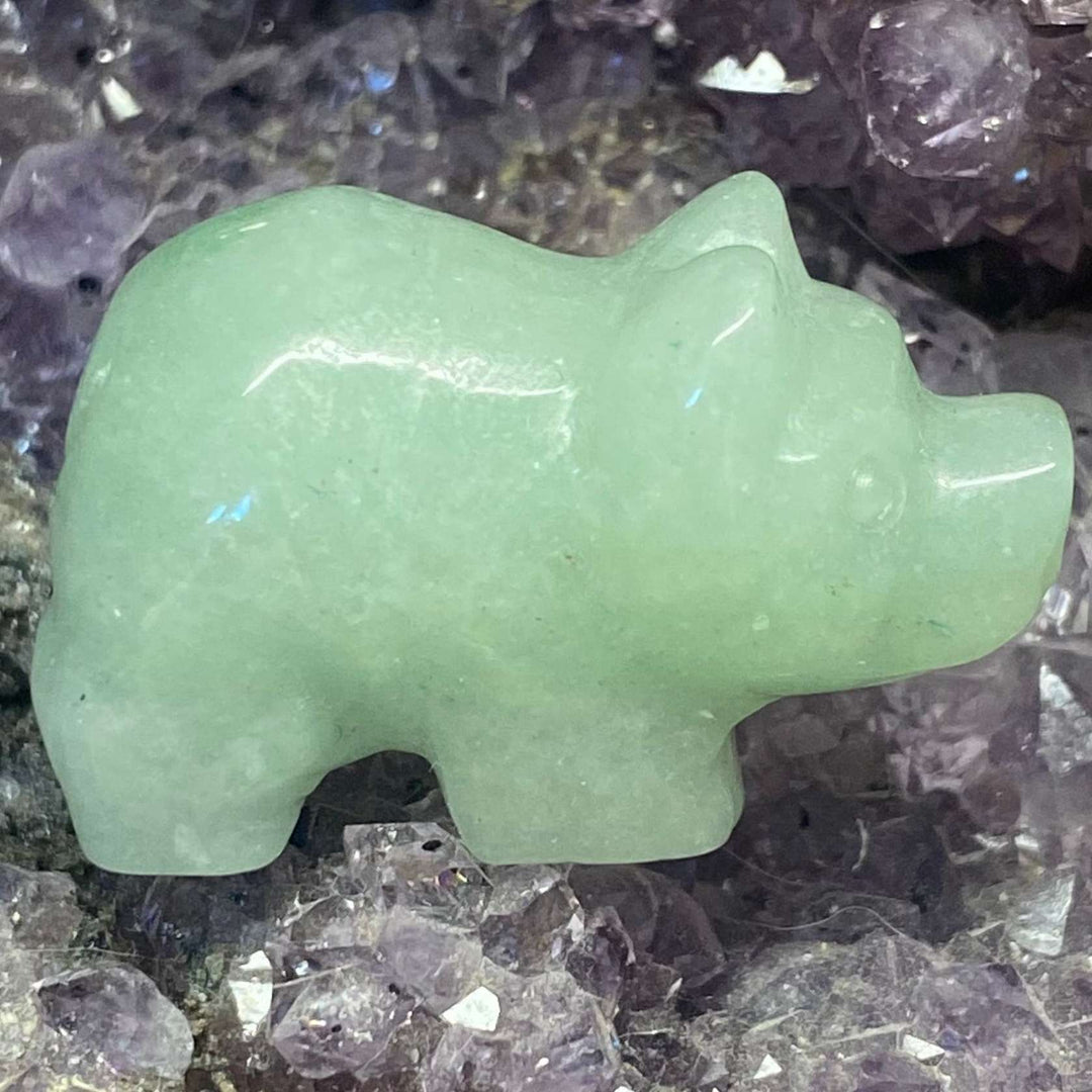 A close-up view of a hand-carved green aventurine pig crystal, showcasing its smooth, silky finish and soft minty-green shading. The 3cm pocket treasure rests on a bed of raw purple amethyst matrix, highlighting its connection to heart chakra energy, soft luck, and steady optimism.